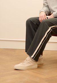 Sneaker beige con tomaia testurizzata, lacci e linguetta imbottita. Indossati con pantaloni verde scuro con strisce bianche sui lati. Sfondo in parquet.