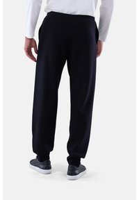 Mann in lockeren schwarzen Hosen und schwarzen Sneakers mit weißen Sohlen, der vor einem schlichten weißen Hintergrund steht.