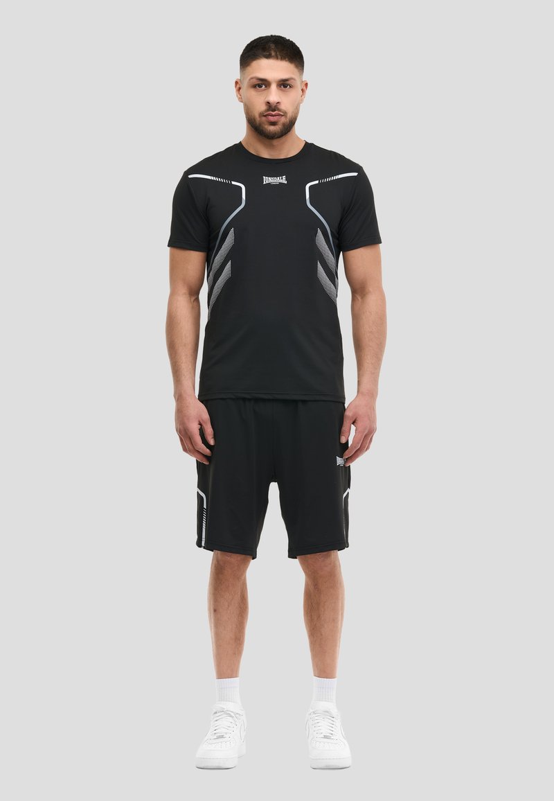 Άνδρας στέκεται φορώντας μαύρο αθλητικό T-shirt και σορτς με λευκές λεπτομέρειες, λευκές κάλτσες και λευκά αθλητικά παπούτσια μπροστά σε γκρι φόντο.
