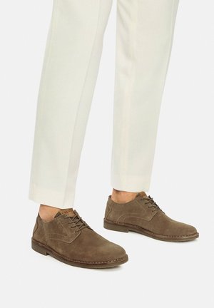 Pánské hnědé semišové boty na šněrování v kombinaci s krémovými společenskými kalhotami, zobrazené od poloviny lýtek dolů na bílém pozadí.