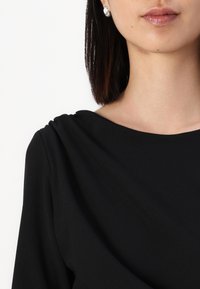 Černý top s hladkou texturou, se zaobleným výstřihem a plisováním na ramenou. Obsahuje kovové náušnice s kamínky, umístěné v úrovni uší.