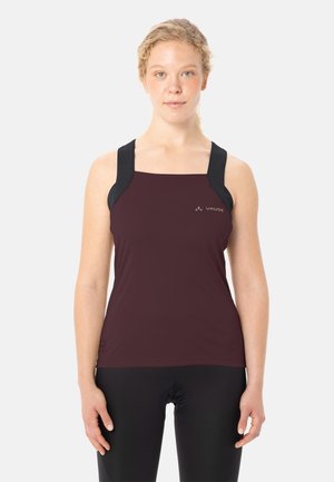 Femme portant un haut de sport bordeaux sans manches avec des bretelles noires et un short de cyclisme noir, debout face à l'avant sur un fond uni.