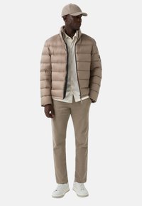 Beige pufferjack met een doorstikt patroon, zwarte rits en hoge kraag, gecombineerd met een licht shirt, beige broek en witte sneakers.