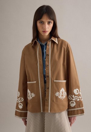 Frau mit glattem braunem Haar trägt eine braune Jacke mit weißer Blumenstickerei über einem blauen Jeanshemd und gestreiften Hosen.