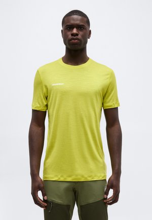 Άνδρας που φοράει φωτεινό κίτρινο κοντομάνικο μπλουζάκι και διχρωματικά πράσινα παντελόνια, στέκεται μπροστά σε απλό ανοιχτό φόντο.