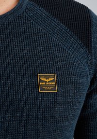 Blaue strukturierte Strickjacke mit einem gerippten Design und einem glatten Schulterbereich. Verfügt über ein quadratisches PME Legend Etikett in Orange und Schwarz.
