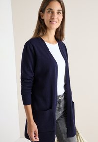Cardigan lungo navy con polsini a costine e due tasche frontali, caratterizzato da una vestibilità rilassata e scollatura a V, indossato sopra un top bianco e jeans grigi.