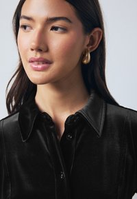 Černá sametová košile s špičatým límcem, se zapínáním na knoflíky. Modelka má na sobě zlaté náušnice ve tvaru slzy a jemný, přirozený make-up.