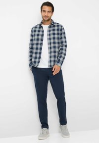 Blauwe en grijze geruite flanellen shirt over een witte t-shirt, gecombineerd met marineblauwe pantalon en lichtgrijze sneakers. Heeft knoopdetails en zakken.