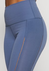Detailní záběr na osobu v modrých legínách s vysokým pasem, které mají žebrované boční panely a malé síťované výřezy podél švů.