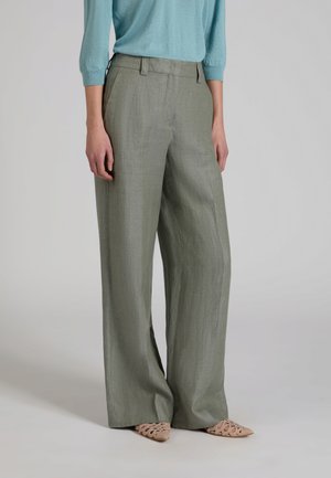 Persona che indossa pantaloni grigi a gamba larga, maglia a maniche lunghe color teal e ballerine beige con tagli e punta appuntita, in piedi su uno sfondo grigio uniforme.