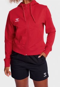 Rode zip-up hoodie met een verstelbare capuchon en zakken aan de voorkant, gecombineerd met zwarte shorts. Beide items hebben logo's en zijn gemaakt van zachte, ademende stof.