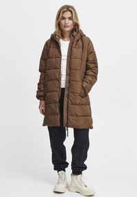 Brun pufferjakke med hætte, der har lynlås foran, quiltet design og snoretræk. Båret over sorte bukser og støvler.