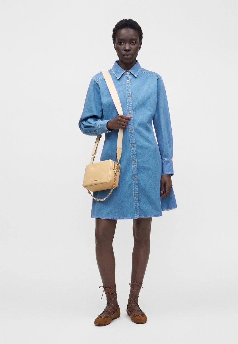 Femme portant une robe-chemise en denim bleu, des ballerines marron à lacets et un sac bandoulière beige avec un détail chaîne.