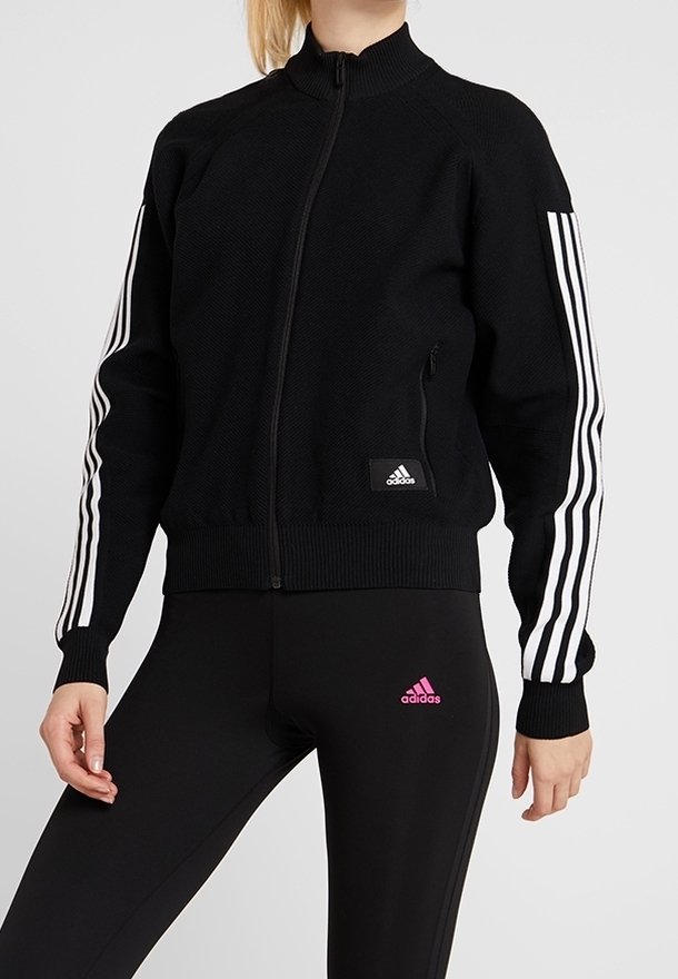 Veste zippée noire avec col montant, tissu texturé, trois bandes blanches sur les manches et petit logo Adidas noir sur le devant.