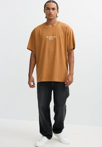 Camiseta de algodón marrón de manga corta, con texto bordado en blanco en la parte delantera, combinada con vaqueros negros y zapatillas blancas.