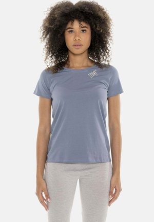 Donna con capelli ricci che indossa una camicia a maniche corte azzurro chiaro e pantaloni grigio chiaro, in piedi di fronte con un’espressione neutra.