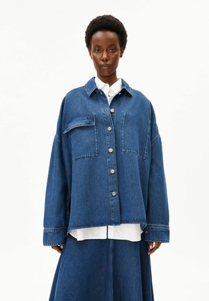 Frau mit kurzem Afrohaar trägt eine übergroße blaue Jeansjacke über einem weißen Hemd und einem passenden blauen Jeansrock, steht vor weißem Hintergrund.