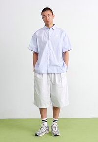 Lyseblå kortermet skjorte med knapper, hvite shorts, grå sneakers og svart-hvite stripe sokker, brukt av en person som står.