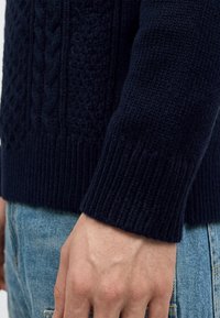 Gros plan d'une main portant une manche de pull tricoté bleu marine foncé avec bord-côtes et un jean en denim bleu.