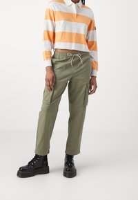 Gestreiftes Polo mit langen orange-weißen Ärmeln, grüne Cargo-Hose mit elastischem Bund und Kordelzug, schwarze Schnür-Stiefel im Combat-Stil.