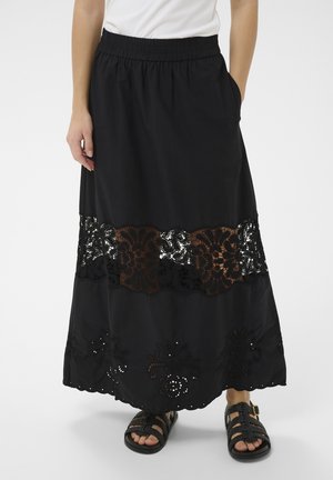 Čierna maxi sukňa so širokým kvetinovým čipkovým panelom a okrajom s očkami, nosená s čiernymi pletenými sandálmi a bielym topom, pohľad spredu orezaný do pása.