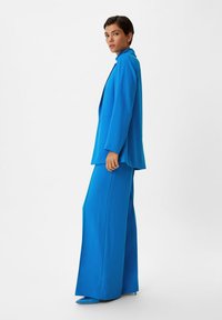 Helderblauwe op maat gemaakte blazer en bijpassende broek met wijde pijpen. De stof lijkt glad. Een slim fit, scherpe revers en puntige schoenen maken de look compleet.