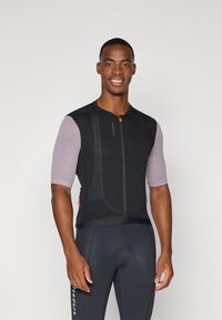 Maillot de cyclisme en mesh noir avec des manches courtes à motifs, fermeture éclair à l'avant et accents orange. Porté par-dessus un short de cyclisme noir.