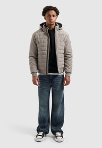 Grijze gewatteerde jas met capuchon, voorrits en geribde mouwen, gecombineerd met donkerblauwe wijde jeans en zwart-witte sneakers.