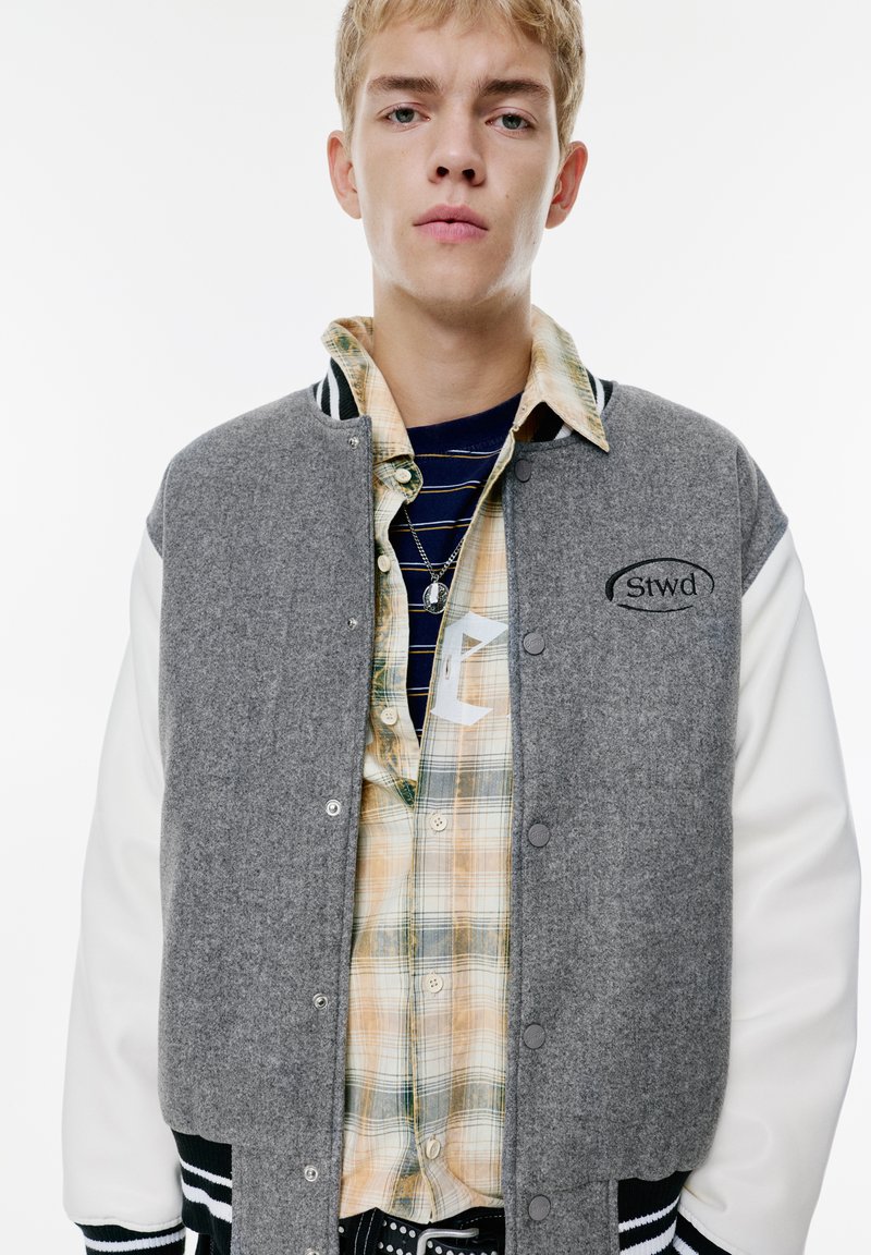 Veste varsity en mélange de laine grise avec des manches blanches et des poignets à rayures noires, portée sur une chemise à carreaux dans des tons de jaune, vert et bleu.