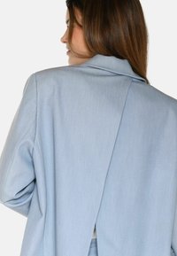 Svetlo modra blazer z prilagojenim krojem. Ima klasičen ovratnik, hrbtno zarezo in gladko teksturo. Narejena je iz tkanine.