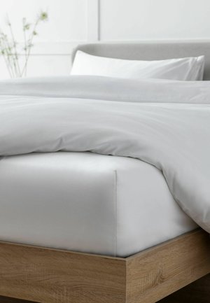 Bett mit weißem Spannbetttuch, weißer Bettdecke und Kissen auf einem Holzrahmen mit grauem Kopfteil und unscharfen Pflanzen im Hintergrund.