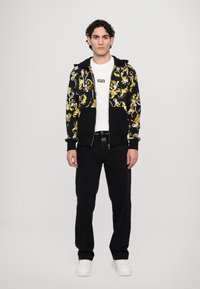 Μαύρο φούτερ με κίτρινο και λευκό λουλούδινο μοτίβο, με φερμουάρ, φορεμένο πάνω από άσπρη μπλούζα και συνδυασμένο με μαύρο παντελόνι και άσπρα αθλητικά παπούτσια.