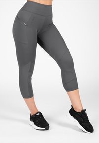 Grijze capri-legging gemaakt van rekbare stof met een hoge tailleband, een ritszak en een gestructureerd detail op het onderbeen. Zwarte sport schoenen inbegrepen.