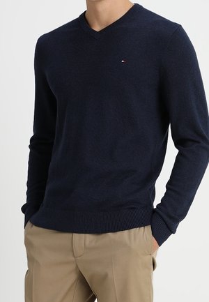 Man draagt een marineblauwe V-hals trui met beige broek, handen in de zakken, tegen een effen witte achtergrond.