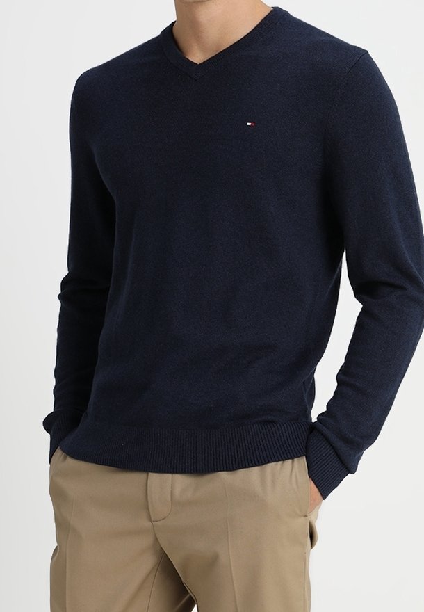 Homme portant un pull bleu marine à col en V avec un pantalon beige, mains dans les poches, sur un fond blanc uni.