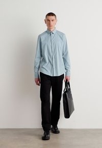 Camisa azul claro de botones con collar puntiagudo, combinada con pantalones y zapatos negros. El modelo sostiene un bolso estructurado negro.