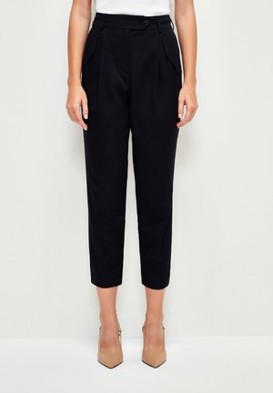 Pantaloni neri sartoriali con vita alta, pieghe frontali e tasche laterali, abbinati a scarpe con tacco beige. Tessuto liscio e taglio dritto.