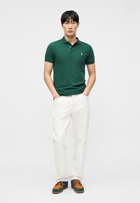 Άνδρας που φοράει σκούρο πράσινο πόλο πουκάμισο, άσπρο παντελόνι και καφέ-πράσινα δετά παπούτσια, στέκεται με τα χέρια στις τσέπες μπροστά σε απλό φόντο.