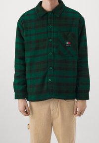 Camicia di flanella a quadri verdi e neri con colletto, bottoni frontali, taschino sul petto e patch con logo sulla tasca. Texture morbida, design casual.