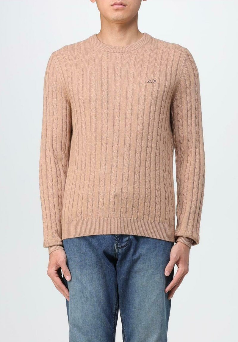 Maglione beige chiaro a maglia cable-knit con collo rotondo, maniche lunghe e polsini e orlo a coste. Piccolo logo sul petto. Abbinato a jeans blu.