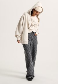 Beige oversized hoodie with drawstring, paired with gray checkered wide-leg pants. The outfit features a cream beanie and black footwear.