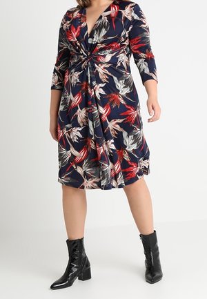 Kvinde iført en marineblå knælange kjole med blomsterprint, trekvart lange ærmer og sorte ankelstøvler med blokhæl.