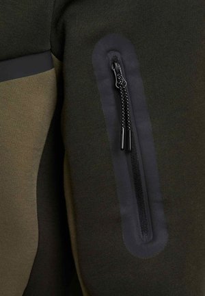 Haut de sport vert et noir avec une poche zippée dotée d'une fermeture éclair noire, texture lisse et panneaux en tissu contrastés sur les côtés.