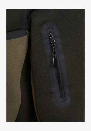 Haut de sport vert et noir avec une poche zippée dotée d'une fermeture éclair noire, texture lisse et panneaux en tissu contrastés sur les côtés.