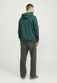 Dunkelgrüner Hoodie mit Kapuze und Kordelzug, kombiniert mit dunkelgrauen Jeans. Glattes Material, lockere Passform, minimales Design.