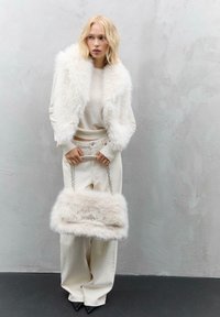 Donna bionda in un gilet di pelliccia sintetica bianca e pantaloni ampi color crema tiene una borsa grande e morbida abbinata con manici a catena contro un muro grigio.