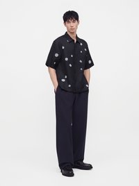 Jeune homme debout, les mains dans les poches, portant une chemise noire à manches courtes avec des fleurs blanches, un pantalon ample foncé et des chaussures noires sur fond blanc.