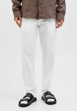 Man draagt witte broek, bruin shirt met lange mouwen, zwarte sandalen en witte sokken, staand tegen een effen achtergrond.