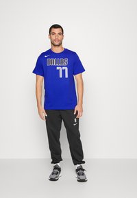 Blå T-skjorte med kort erme som har "DALLAS" og "77" trykt i hvitt og grått, sammen med svarte joggebukser og sportssko.